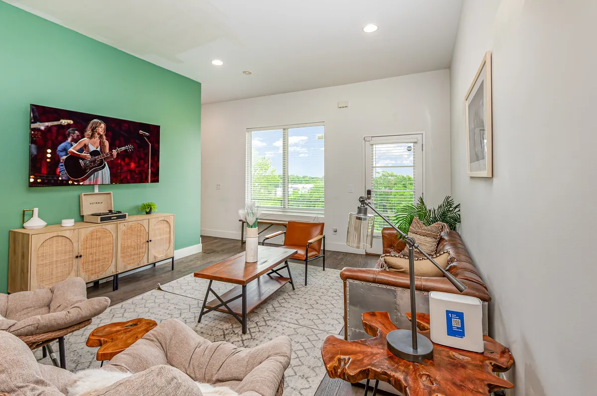 A spacious and contemporary living room featuring a large TV displaying a musical performance, a green accent wall, stylish coffee table, and a comfortable seating arrangement. Sunlight streams in through the windows, enhancing the room's welcoming atmosphere.
