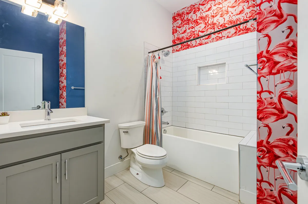 A cheerful bathroom decorated with playful flamingo-patterned wallpaper, a sleek modern vanity, large mirror, and a bathtub with a shower. The space feels bright and welcoming, ideal for guests needing a refreshing start or end to the day.