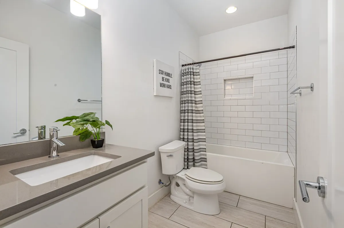 A cozy guest bathroom featuring a traditional vanity with a sink, a toilet, a bathtub with a striped shower curtain, and a touch of greenery from a potted plant. The design is inviting and functional, ideal for family or group stays.