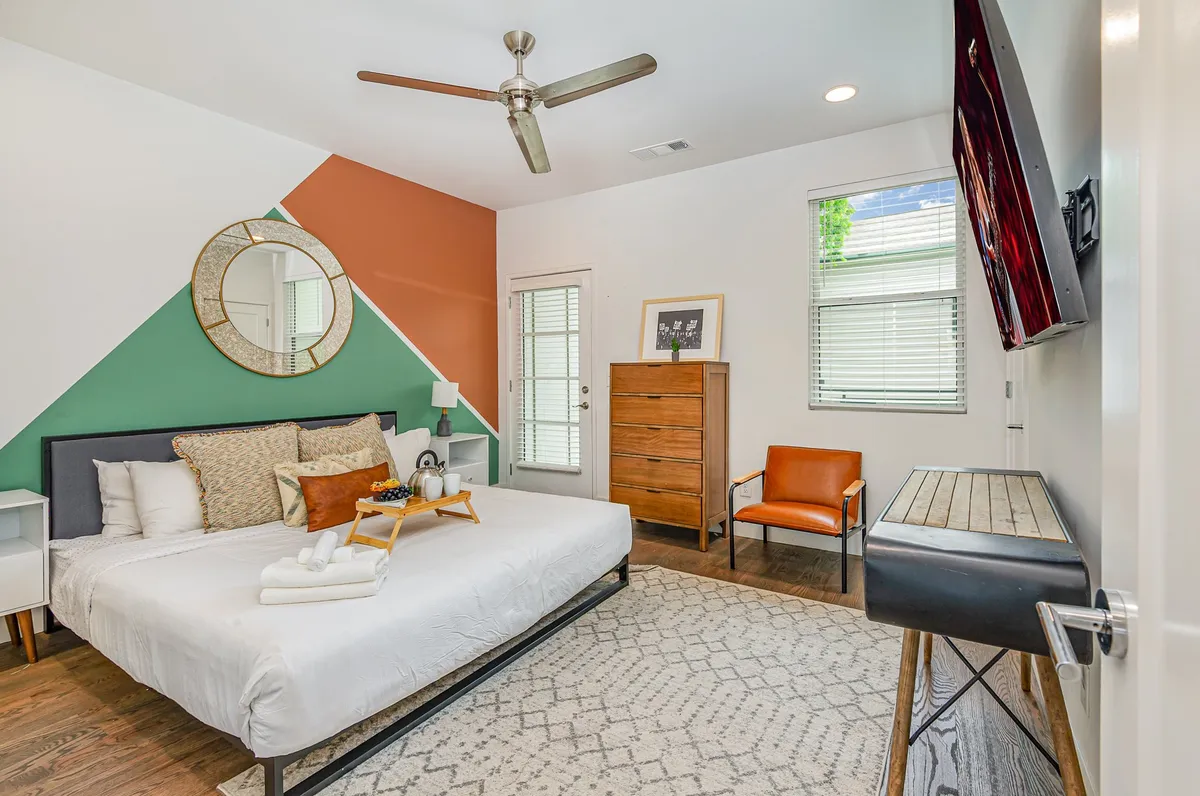 A stylish and comfortable king bedroom featuring a plush bed with decorative pillows, a wall-mounted TV, a mirror, an accent chair, and a dresser. Ample natural light fills the space, giving it a cozy yet refined feel perfect for relaxation.