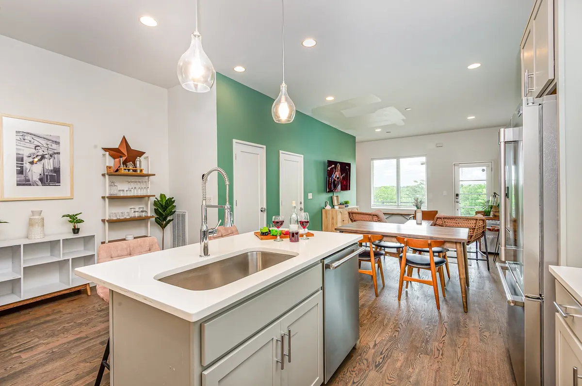 A warm and inviting kitchen and dining space featuring an island with seating, stainless steel appliances, a dining table, a green accent wall, and decorative touches like plants. The layout is open and encourages socializing.