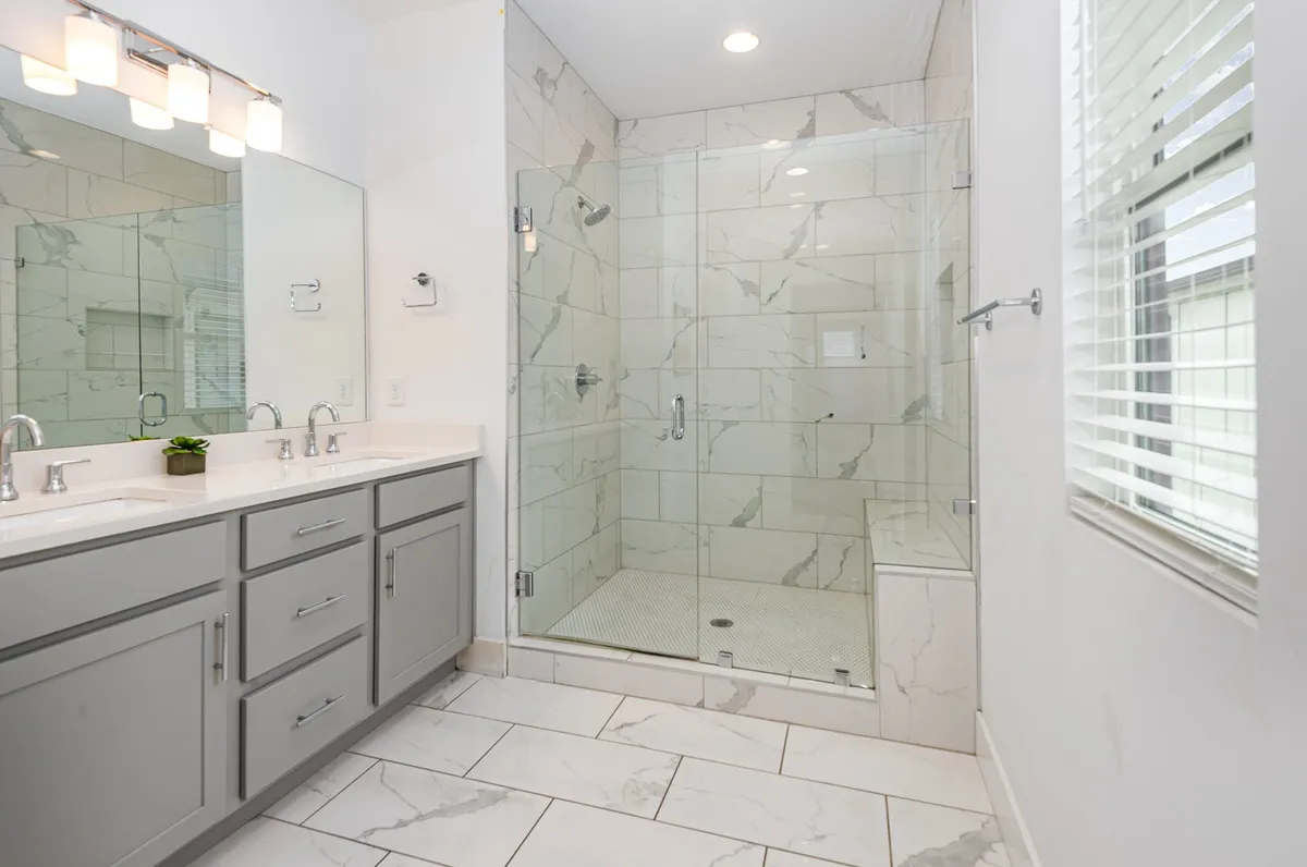 This image showcases a modern bathroom with a double sink vanity, large mirrors, and a glass-enclosed stand-in shower set against elegant marble tile flooring. The space is filled with natural light and has a clean, spa-like aesthetic.