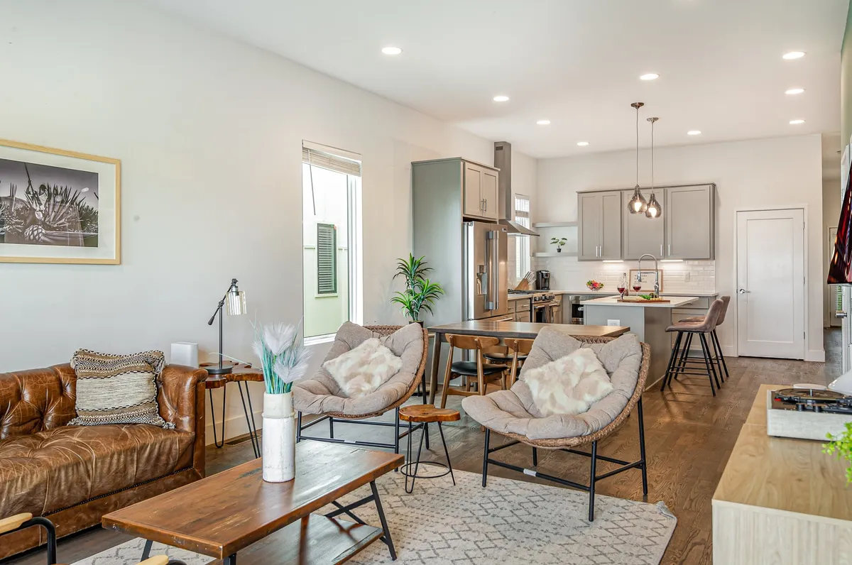 The image showcases a bright, open-concept living space with a plush leather sofa, a spacious dining table, a sleek kitchen island with modern hanging light fixtures, and touches of greenery from house plants. The layout invites relaxation, entertaining, and shared meals, all set in a comfortable and stylish environment.